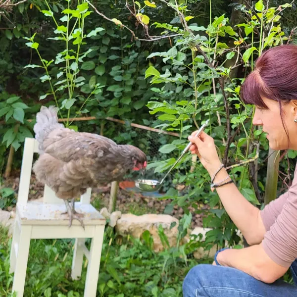 Huhn sitzt auf einem kleinen Stuhl, daneben eine Frau im Garten